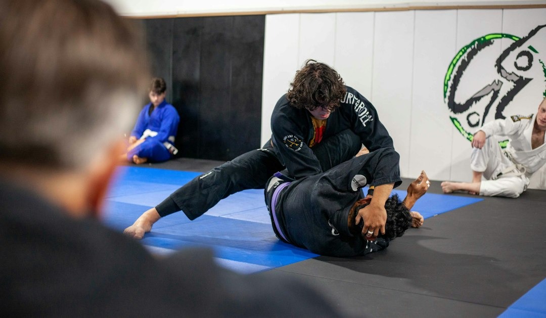 Two people wearing black gis grapple on gym mats.